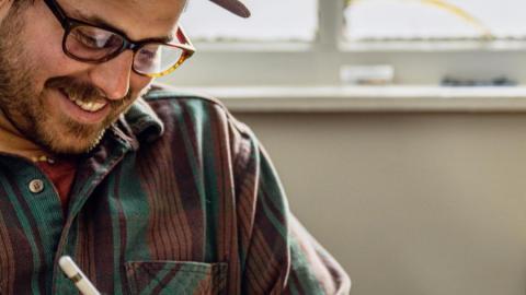 Image of the artist wearing a flannel and hat and drawing on a tablet.